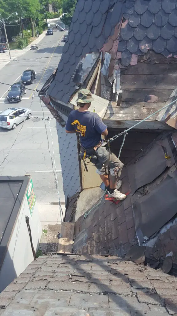 toronto roofer in a harness on the side of a roofing turrett on a very steep roof providing a roof repair by replacing roof shingles in toronto