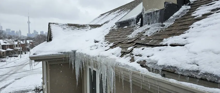 Severe ice dam damage with large icicles hanging from eavestrough and snow covered shingles on a Toronto home roof in winter with CN Tower visible in background &mdash; Right Choice Roofing and Repair