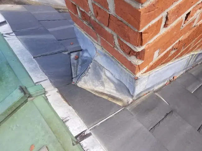 Progress photo showing wildlife damaged and deteriorating lead flashing cricket at base of brick chimney on slate roof in Harbord Village Toronto prior to copper conversion by Right Choice Roofing and Repair