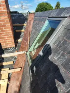 Local Toronto slate roofer's view looking down slope showing Velux skylight with copper flashing being fitted and new slate tiles installed on either side alongside brick chimney during slate roof replacement in Harbord Village Toronto by Right Choice Roofing and Repair